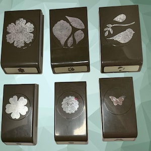 May include: A set of six dark brown craft paper punches arranged in two rows. The punches feature various die-cut shapes including a flower, leaves, a bird on a branch, a scalloped circle with 1 3/4 inch text, and a butterfly design.