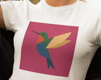 Puede incluir: Una camiseta blanca con un diseño de colibrí geométrico impreso en la parte delantera. El colibrí es verde, azul, amarillo y naranja sobre un fondo rosa.