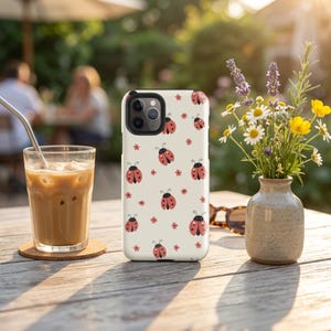 K&ouml;nnte beinhalten: Wei&szlig;es iPhone-Case mit einem sich wiederholenden Muster aus roten Marienk&auml;fern und kleinen roten Blumen. Das Case liegt auf einem Holztisch neben einem Glas Eiskaffee und einer kleinen Blumenvase.