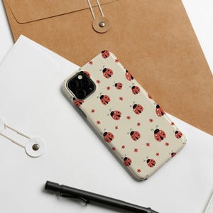 K&ouml;nnte beinhalten: Eine beige Handyh&uuml;lle mit einem sich wiederholenden Muster aus roten Marienk&auml;fern und kleinen roten Blumen. Die H&uuml;lle liegt auf einer wei&szlig;en Oberfl&auml;che, neben einem schwarzen Stift, einer B&uuml;roklammer und einem braunen Umschlag.