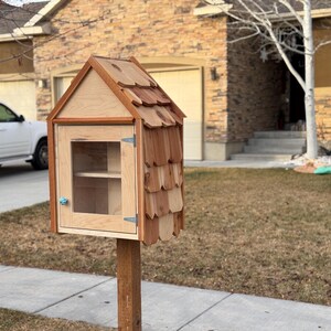 May include: A wooden little free library with a shingled roof, a glass door, and two shelves inside. The library is mounted on a wooden post.
