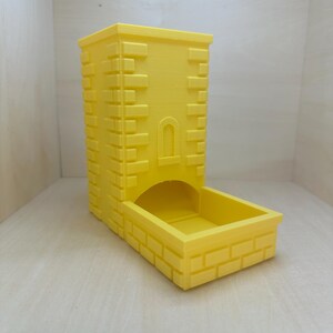 May include: A yellow 3D printed dice tower with a brick pattern. The tower has a small archway opening at the top and a rectangular base.