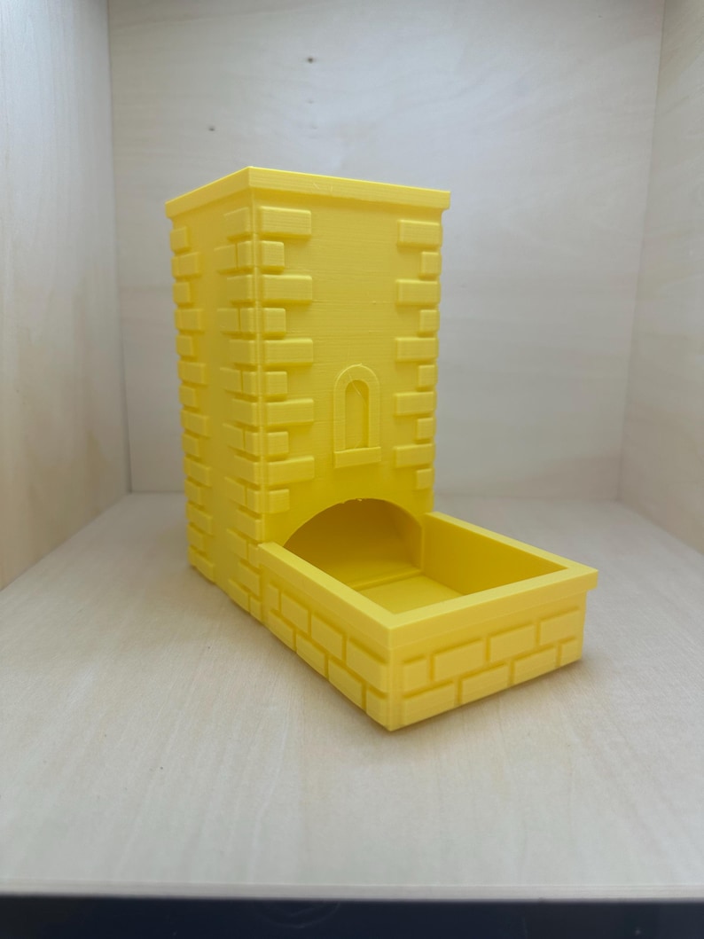 May include: A yellow 3D printed dice tower with a brick pattern. The tower has a small archway opening at the top and a rectangular base.