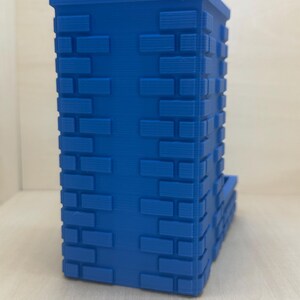May include: A blue 3D printed dice tower with a brick pattern. The tower has a slot for dice to roll out of.