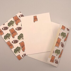May include: Three white note cards with a brown outline of the state of Alabama and the word "Alabama" written inside. The note cards have a brown and white pattern of footballs and trees.