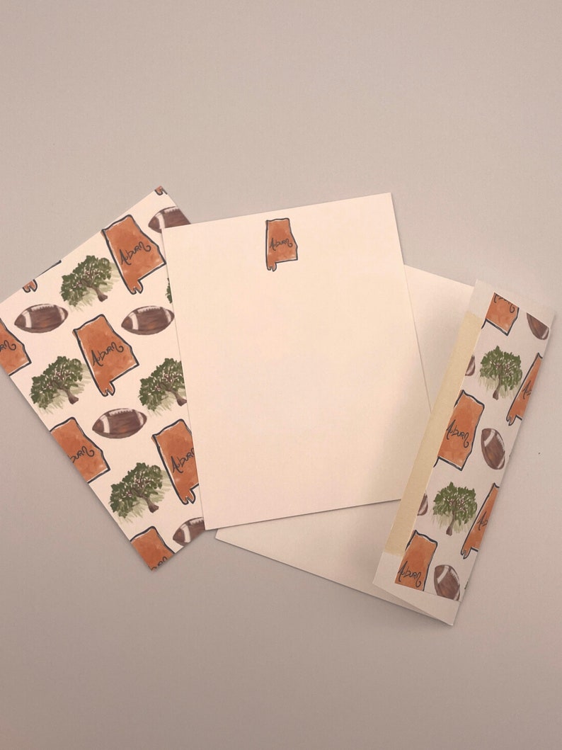 May include: Three white note cards with a brown outline of the state of Alabama and the word "Alabama" written inside. The note cards have a brown and white pattern of footballs and trees.