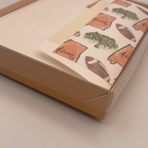 May include: A box of Auburn University themed stationery with a white background and a repeating pattern of orange outlines of the state of Alabama, green trees, and brown footballs. The text "Auburn" is written in cursive on the paper.