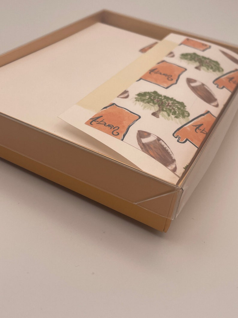 May include: A box of Auburn University themed stationery with a white background and a repeating pattern of orange outlines of the state of Alabama, green trees, and brown footballs. The text "Auburn" is written in cursive on the paper.