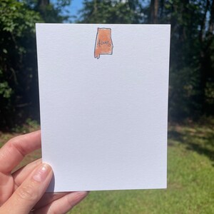 May include: A white card with a brown outline of the state of Alabama with the word "Alabama" written inside. The card is held in a hand against a green grassy background.
