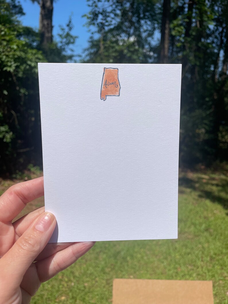 May include: A white card with a brown outline of the state of Alabama with the word "Alabama" written inside. The card is held in a hand against a green grassy background.