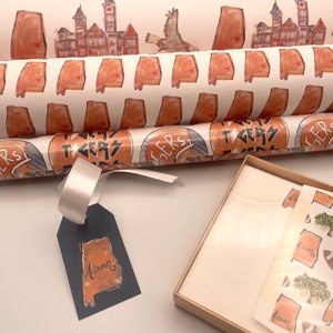 May include: Two rolls of wrapping paper with a white background and orange and brown designs. The top roll features a repeating pattern of the state of Alabama. The bottom roll features a repeating pattern of a circle with the words "Roll Tide" and "Tigers". A gift tag with a blue background and an orange and brown outline of the state of Alabama with the word "Auburn" written inside. A small gift box with a white background and a brown border. The box contains a sheet of wrapping paper with a repeating pattern of the state of Alabama, a tree, and a football.