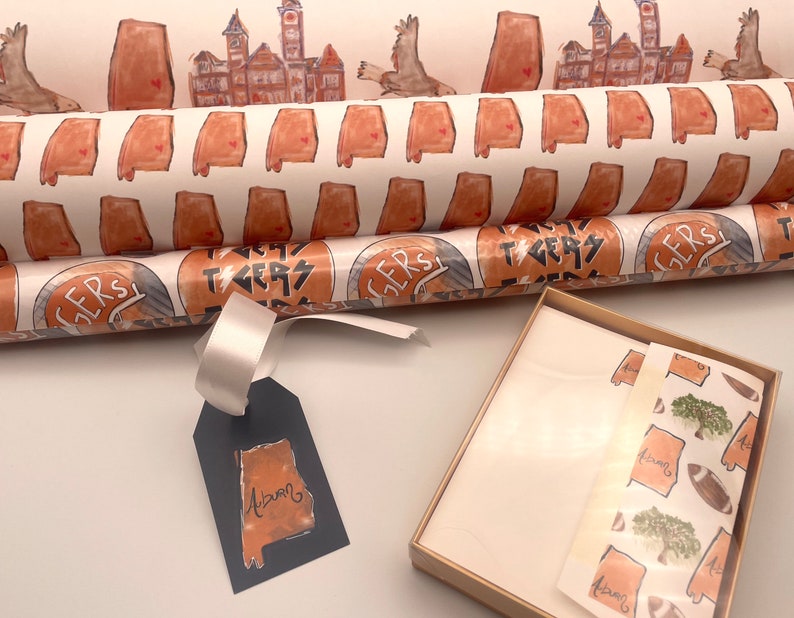 May include: Two rolls of wrapping paper with a white background and orange and brown designs. The top roll features a repeating pattern of the state of Alabama. The bottom roll features a repeating pattern of a circle with the words "Roll Tide" and "Tigers". A gift tag with a blue background and an orange and brown outline of the state of Alabama with the word "Auburn" written inside. A small gift box with a white background and a brown border. The box contains a sheet of wrapping paper with a repeating pattern of the state of Alabama, a tree, and a football.
