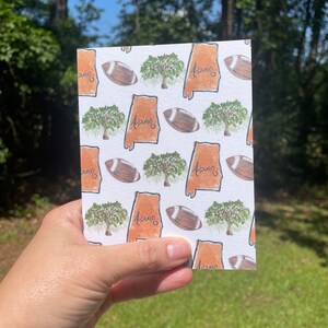 May include: A white card with a repeating pattern of an orange outline of the state of Alabama, a green tree, and a brown football. The text "Auburn" is written in black inside the state outline.