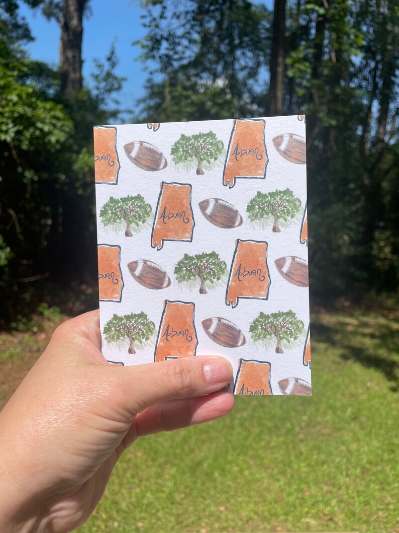 May include: A white card with a repeating pattern of an orange outline of the state of Alabama, a green tree, and a brown football. The text "Auburn" is written in black inside the state outline.