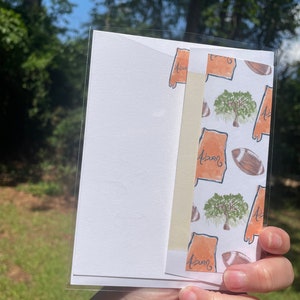 May include: A white card with a cream-colored envelope attached. The envelope has a pattern of orange outlines of the state of Alabama with the word "Auburn" written inside each outline. There are also images of trees and footballs.