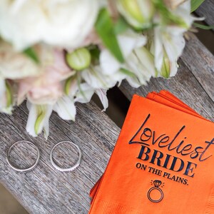 May include: A white bridal bouquet with two silver wedding bands on a wooden surface. A bright orange napkin with the text "Loveliest Bride on the Plains." is in the background.
