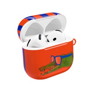 May include: Open orange AirPods case with a blue and red striped top. The case features a cartoon alligator wearing blue glasses. Two white AirPods are inside the case.