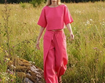 May include: A bright pink two-piece set consisting of a short-sleeved, boxy crop top and matching wide-leg trousers. The trousers feature a distinctive wrap-style front panel with a tie detail at the waist, creating a layered appearance.
