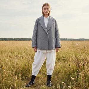 Könnte beinhalten: Eine Person trägt eine grau gesteppte Jacke, ein weißes Hemd und eine weiße Hose. Die Jacke hat einen klassischen Kragen und zwei Taschen. Sie trägt schwarze Stiefel und steht in einem Feld mit hohem Gras.