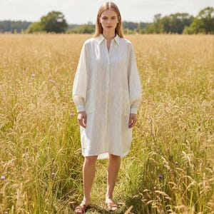 Könnte beinhalten: Weißes Kleid mit vertikalen schwarzen Streifen. Es hat einen Hemdkragen, eine Knopfleiste und zwei Vordertaschen. Das Model trägt braune Sandalen. Der Hintergrund ist ein Feld mit hohem Gras.
