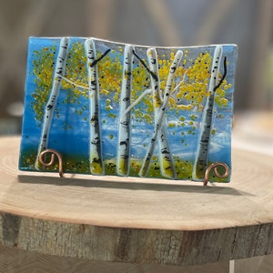 May include: A glass art piece depicting a scene of birch trees with white bark and yellow leaves against a blue sky background. The piece is mounted on a wooden stand with copper wire supports.