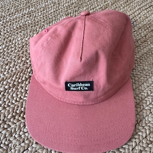 May include: A dusky pink baseball cap with a curved peak. The cap has the text "Caribbean Surf Co." on a small black rectangular patch. The cap is made of a durable fabric and has a button on top.