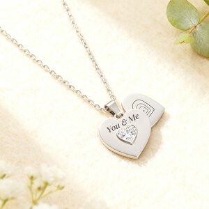 Op de afbeelding: Een zilverkleurige hartvormige hangerketting met de woorden "You & Me" en een helder kristallen hart. De ketting heeft een delicate ketting en een tweede hartvormige bedel met een geëtst ontwerp. Een romantisch sieraad.