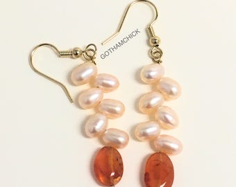 May include: A pair of gold dangle earrings with a cascade of white pearls and a single orange carnelian stone at the bottom.  The earrings are marked "GOTHAMCHICK".