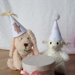 Könnte beinhalten: Zwei Stofftiere, ein Hund und ein Lamm, tragen Partyhüte und sitzen neben einer runden Geschenkbox, die mit einem rosa Band gebunden ist. Der Hut des Hundes hat eine Bärenillustration, der des Lamms hat Punkte.