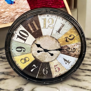 May include: A round, black metal clock with a distressed, vintage-style face. The clock face is divided into 12 sections, each with a different colour and number. The clock hands are black and the numbers are in a white, yellow, and red font.