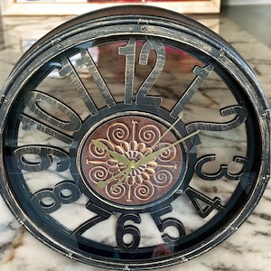 May include: A round clock with Roman numerals and a decorative centre. The clock is black and gold with a distressed finish and a glass face.