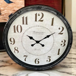 May include: A round clock with a black and silver finish. The clock face is white with Roman numeral markings and the text "Westminster Clock Company" and "London".