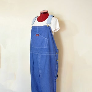 Può includere: Salopette di jeans blu con una maglietta bianca sotto. La salopette ha un'ampia tasca frontale, spalline regolabili e finiture color ottone. Un piccolo logo rosso è visibile sulla tasca frontale. La salopette è esposta su un manichino.