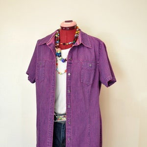 May include: A purple denim shirt dress with short sleeves, button-up front, and two chest pockets. The dress is layered over a white t-shirt and denim shorts. A colorful beaded necklace and patterned belt complete the outfit.