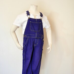 May include: A pair of indigo blue denim overalls with a chest pocket and brass-colored hardware. The overalls are paired with a white short-sleeved shirt. The stitching is a contrasting yellow color.