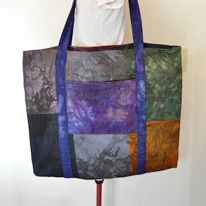 May include: Reversible tote bag with a patchwork design featuring shades of purple, blue, green, and brown. The bag has two long straps and a large main compartment.