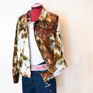 May include: A tie-dye denim jacket in shades of brown, white, and green is displayed on a mannequin. The jacket is paired with a white tank top, denim shorts, and a colorful belt. A beaded necklace adds a pop of color.