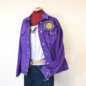 May include: A purple corduroy jacket with vertical stripes, worn over a white tank top and denim shorts. The outfit includes a floral belt, beaded necklaces, and a colorful circular brooch. The mannequin is on a red stand.