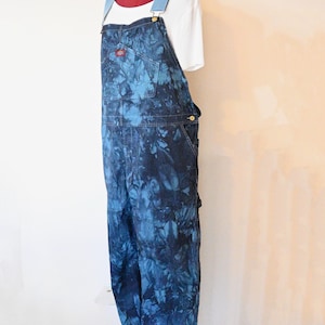 Può includere: Salopette in denim blu tie-dye con spalline regolabili e più tasche. Sotto è indossata una maglietta bianca. Il logo Dickies è visibile sulla tasca anteriore. Lo stile generale è casual. La salopette è su un manichino.