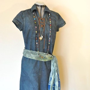 Op de afbeelding: Een spijkerjurk met korte mouwen en een kraag, gepresenteerd op een mannequin. De jurk heeft een veterdetail bij de borst en is geaccessoireerd met een kralenketting, een groene sjaal en een riem. De jurk is ongeveer 91 cm lang.