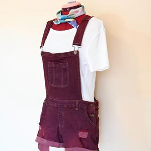 Könnte beinhalten: Ein Outfit mit burgunderfarbenen Jeans-Latzshorts auf einer Schaufensterpuppe. Die Shorts haben Distressed-Details und umgeschlagene Manschetten. Ein weißes T-Shirt und ein farbenfrohes Halstuch vervollständigen den Look. Das Outfit wird auf einem roten Ständer präsentiert.