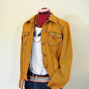 Puede incluir: Chaqueta vaquera color mostaza con botones y detalles de bolsillo, superpuesta a una camiseta blanca y pantalones cortos vaqueros. Un collar de cuentas y un cinturón estampado completan el conjunto. La cabeza del maniquí está sobre una forma roja.