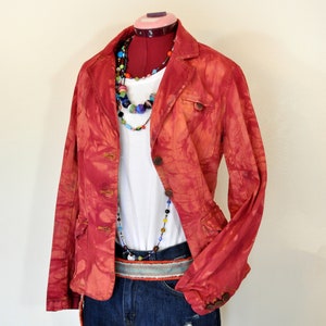 Op de afbeelding: Een rode tie-dye blazer met een wit t-shirt, een spijkerbroek en een kleurrijke kralenketting. De outfit is gecombineerd met een riem met patroon. De blazer heeft knoopsluitingen en zakken.