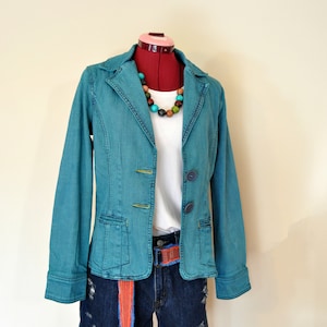 May include: A teal denim jacket with two buttons, worn with distressed denim shorts and a white t-shirt. A colourful beaded necklace and a belt with orange and blue accents complete the outfit. The outfit is displayed on a red dress form.