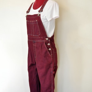 Op de afbeelding: Bordeaux denim overall met verstelbare bandjes en meerdere zakken. De overall wordt gecombineerd met een wit t-shirt. Het kledingstuk wordt gepresenteerd op een mannequin tegen een effen witte achtergrond. De overall heeft knoopsluitingen.