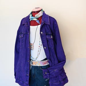 Könnte beinhalten: Auf einer Schaufensterpuppe wird ein Outfit präsentiert: eine lila Jeansjacke, ein weißes Tanktop und Jeansshorts. Dazu gehören ein farbenfrohes Halstuch, Perlenketten und ein floraler Gürtel. Der Look ist für den Alltag gedacht.