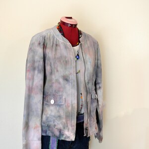 May include: A tie-dye jacket in shades of gray, pink, and blue, with a zipper closure and two front pockets. The jacket is paired with a white top, denim shorts, and a beaded necklace. The outfit is displayed on a mannequin.