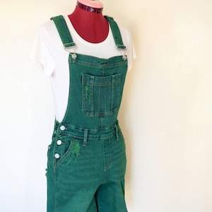 Op de afbeelding: Groene denim overall shorts met distressed details, weergegeven op een mannequin. De shorts hebben verstelbare bandjes, zakken en knoopsluitingen. Er wordt een wit t-shirt onder gedragen.