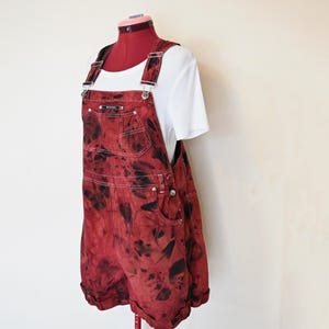 Könnte beinhalten: Rote und schwarze Jeans-Latzshorts mit Batikmuster, verstellbaren Trägern und Taschen. Die Shorts werden mit einem weißen T-Shirt kombiniert und auf einer Schaufensterpuppe präsentiert. Die Latzshorts haben ein einzigartiges, auffälliges Design.