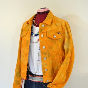 Puede incluir: Chaqueta vaquera de color amarillo mostaza con botones plateados, bolsillos en el pecho y botones. La chaqueta se combina con una camiseta blanca, pantalones cortos vaqueros y un collar de cuentas de colores. También se ve un cinturón rojo y blanco.